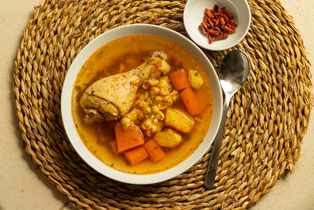 A comforting chicken and vegetable soup served in a bowl with a spoon on a woven mat.