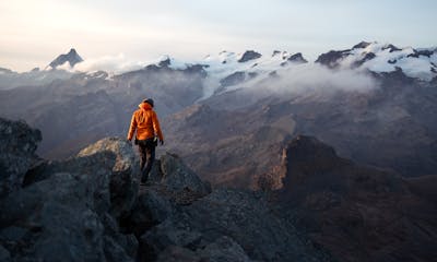 A lone hiker in an orange jacket explores a stunning mountain landscape at dawn, surrounded by misty peaks.