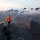 A lone hiker in an orange jacket explores a stunning mountain landscape at dawn, surrounded by misty peaks.