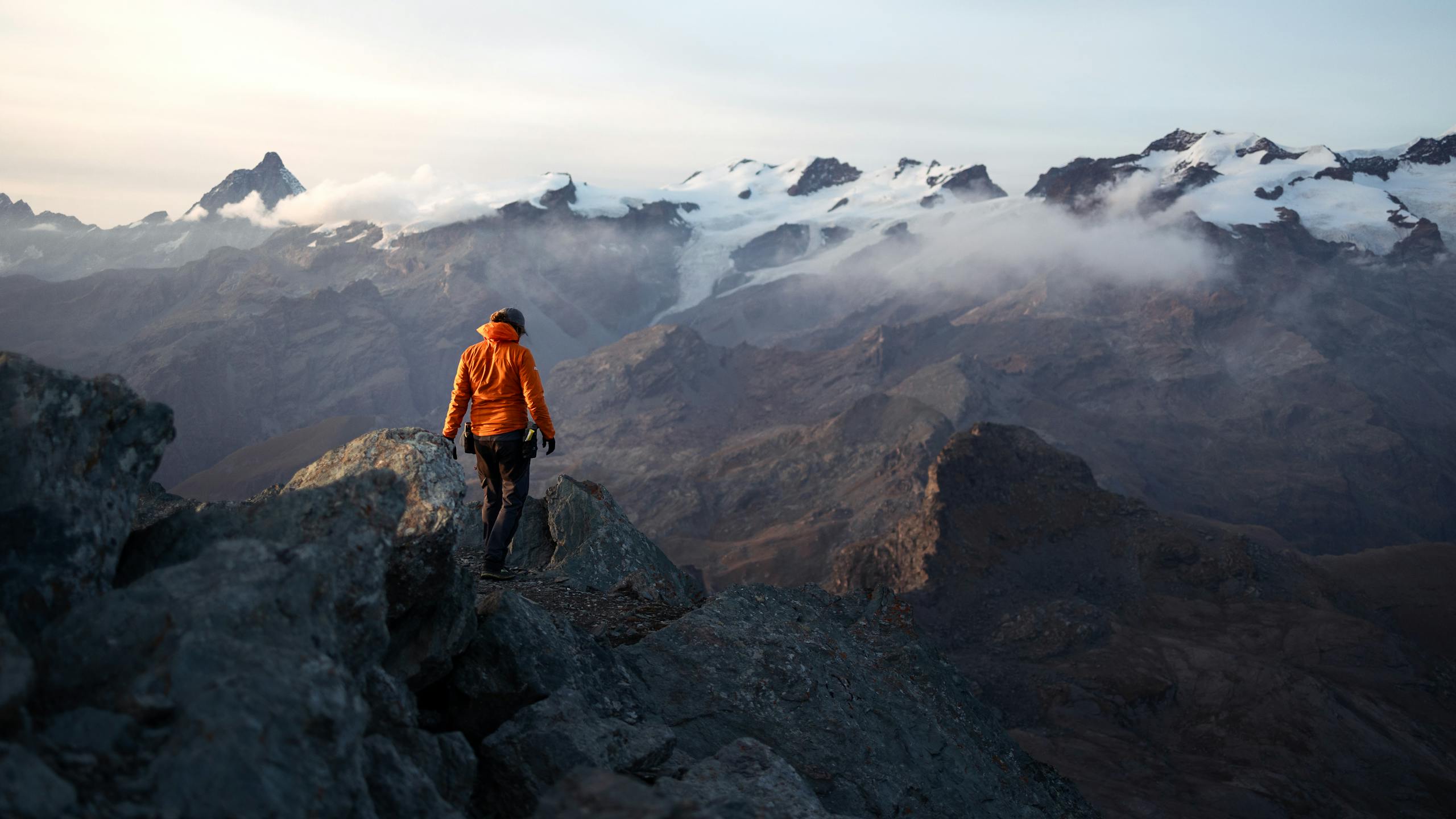 A lone hiker in an orange jacket explores a stunning mountain landscape at dawn, surrounded by misty peaks.