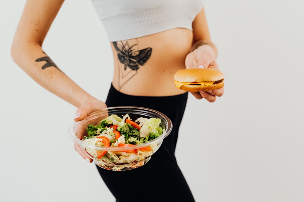 A woman with a tattoo holding a healthy vegetable salad and a cheeseburger, representing food choices.