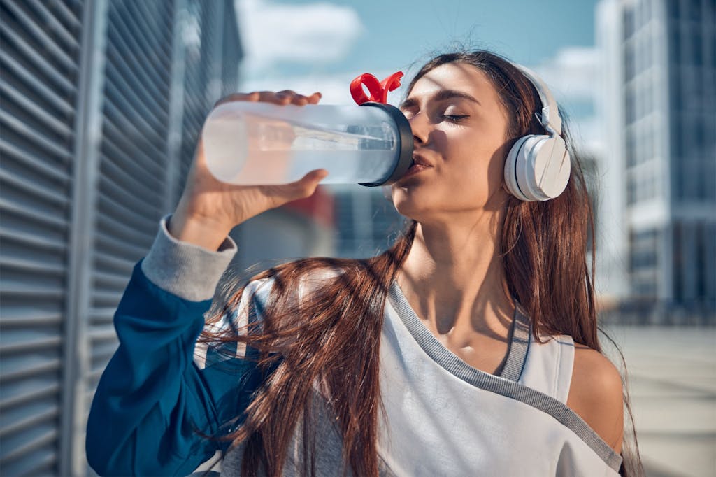 Young woman with headphones enjoying water after workout. Outdoor city background, sunny day.